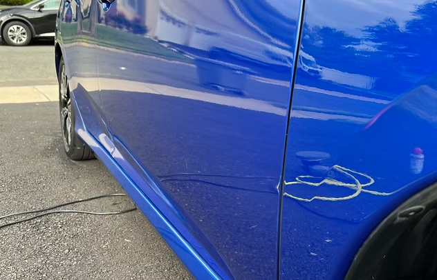 Close-up of a blue car’s side panel with a noticeable dent and scratch near the rear wheel.
