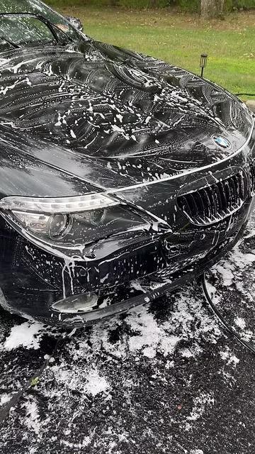 Black sedan covered in white foam from washing, parked on a wet driveway.