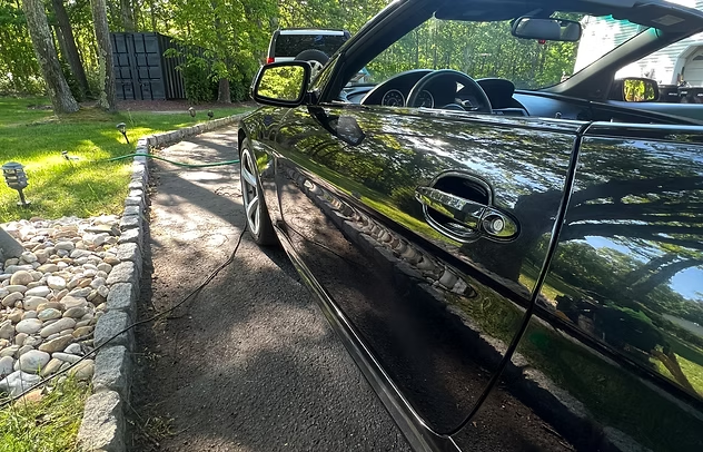 Black car parked in a driveway beside a stone border and green lawn, reflecting trees and sunlight