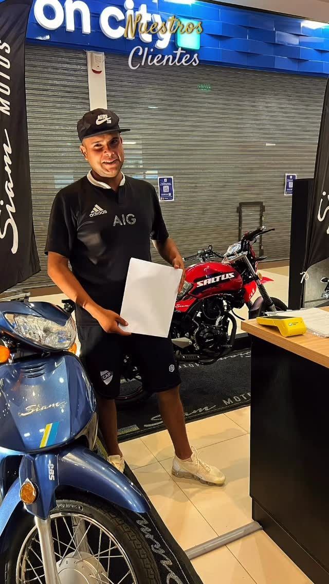 Una persona sonriente con uniforme negro sostiene un papel junto a motocicletas azules y rojas en una sala de exposición.