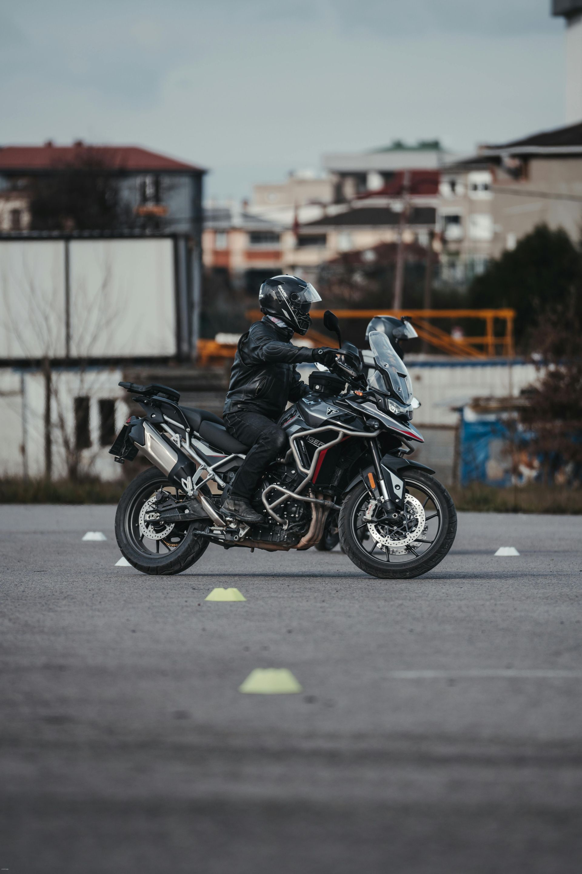 Motociclista en una moto deportiva negra en un estacionamiento, visto de frente con edificios borrosos al fondo.