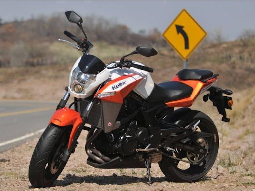 Motocicleta deportiva naranja y negra estacionada en una carretera junto a una señal amarilla de giro a la izquierda.