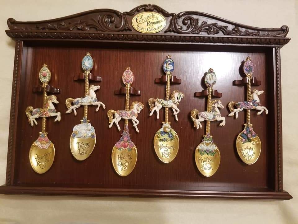 A wooden display with seven decorative carousel spoons, each with a different horse and color.