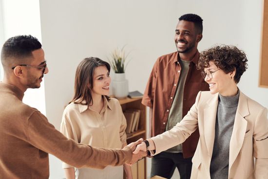 Four coworkers in a bright office shake hands while two others smile nearby