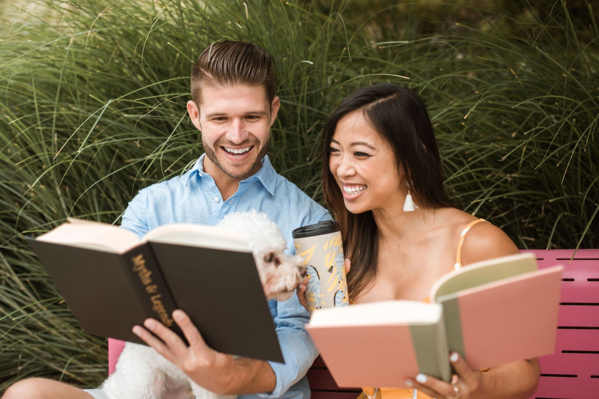 Two people smiling and reading books together outdoors in tall grass
