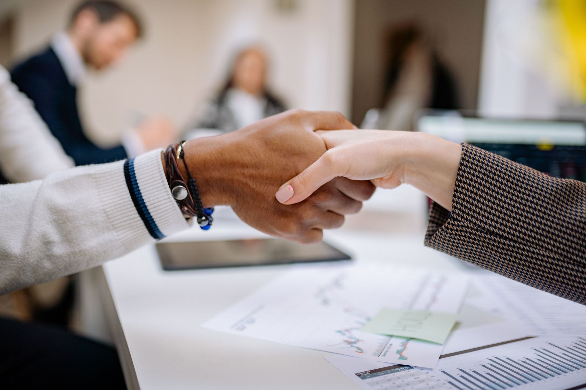 Two people shaking hands over a desk with papers in a meeting room