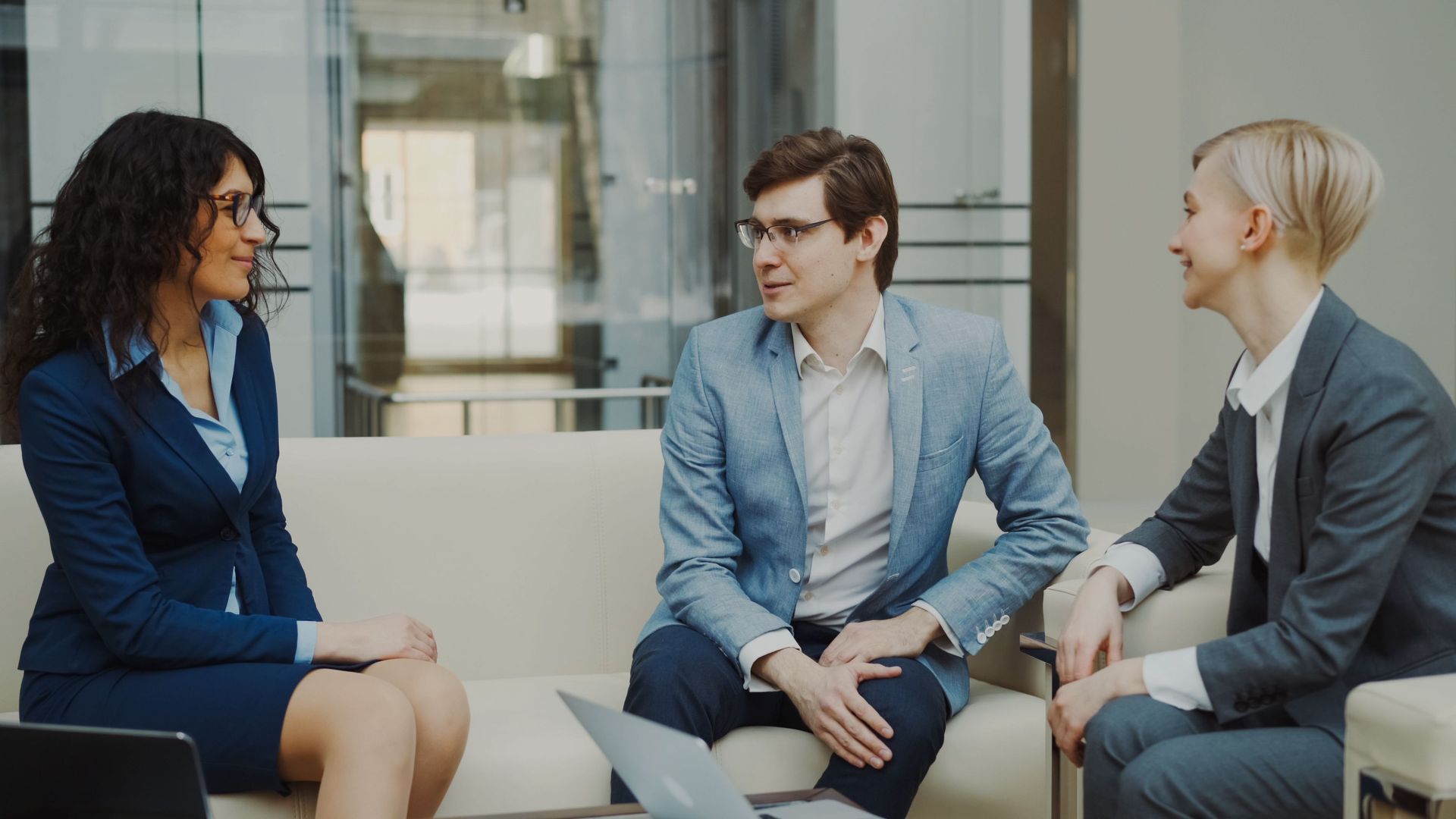 Three coworkers in business attire are seated in a bright office, talking in a meeting.