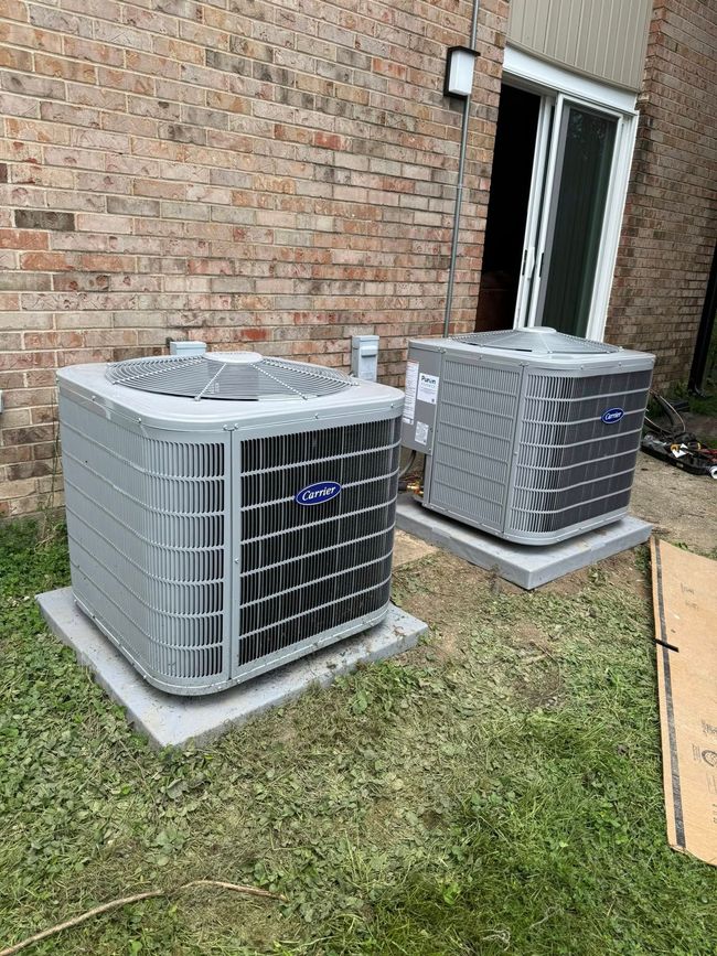 Two gray outdoor air conditioners beside a brick house, one near an open doorway.