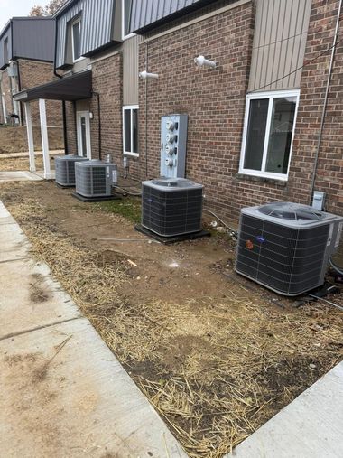 Several outdoor air conditioning units along a brick apartment building beside a sidewalk and mulch bed