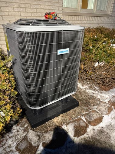 Outdoor air conditioner unit beside a house, surrounded by snow and shrubs.