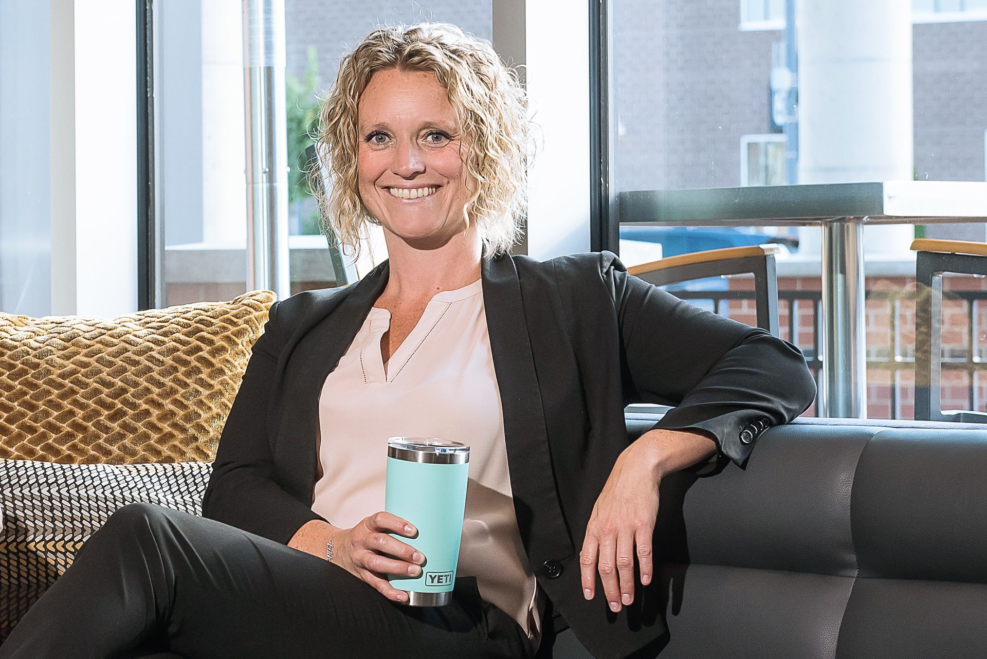 Woman sitting on a couch, smiling, holding a blue tumbler. Wearing a blazer, light-colored blouse, and dark pants. Interior setting.