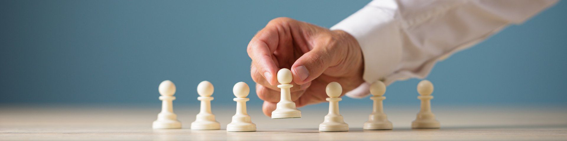 A hand in a white shirt moves a white chess pawn on a wooden surface against a blue backdrop.