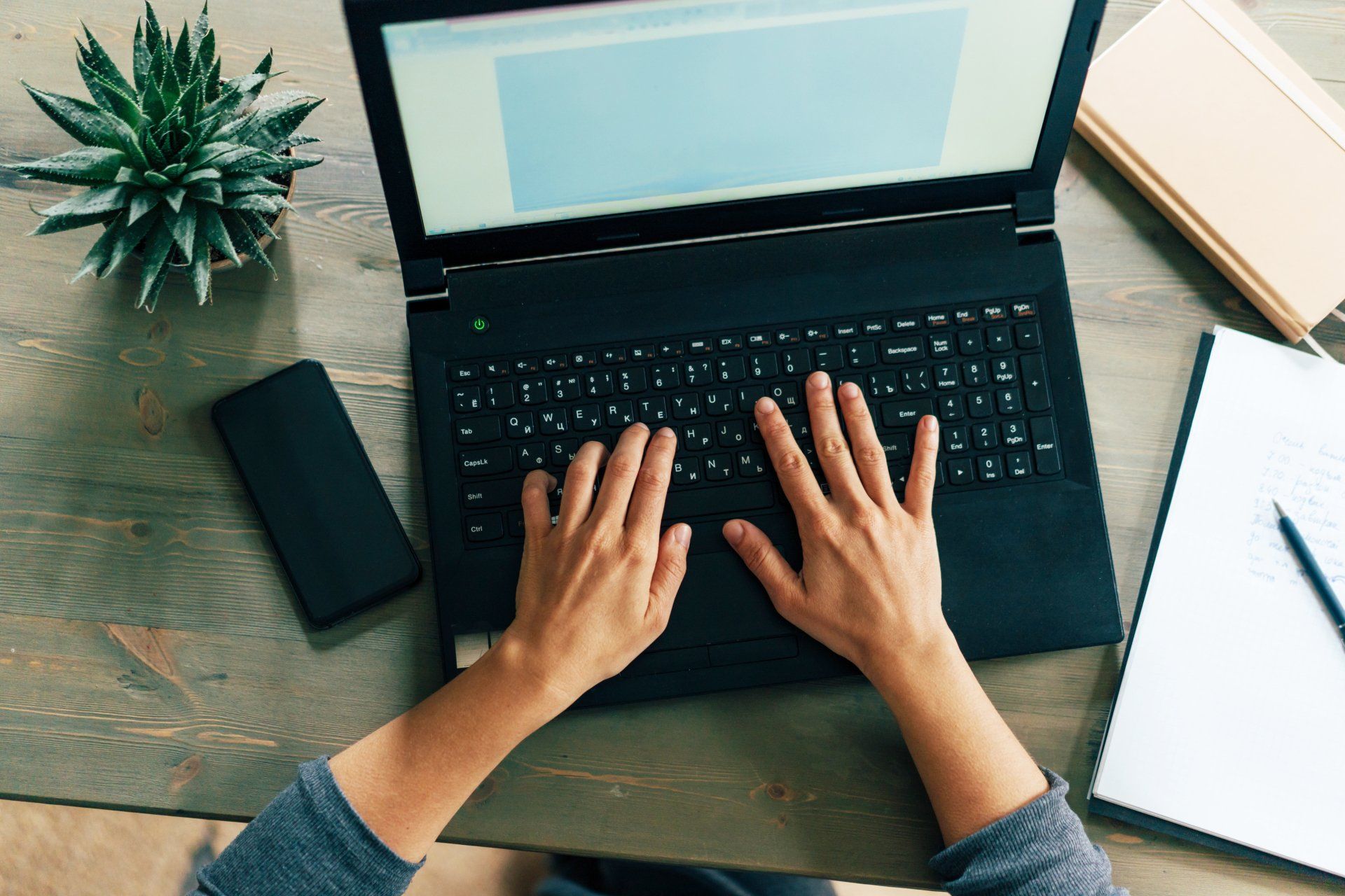 Hands typing on a black laptop, sitting at a wooden desk with a phone, succulent, notepad, and books.