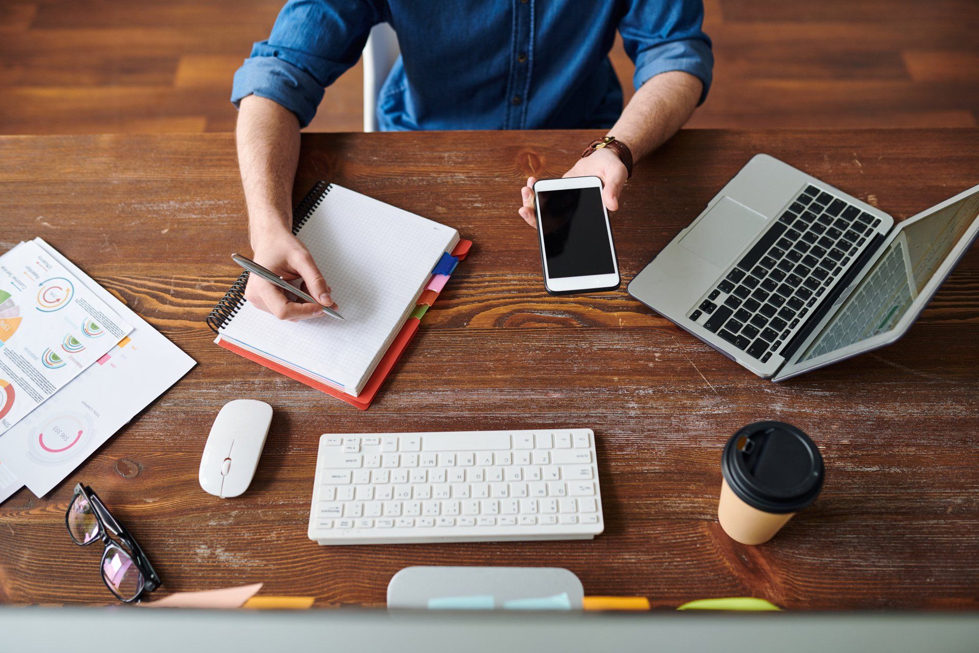 Person writing in notebook, holding phone, and using a laptop, on a wooden desk with a keyboard, coffee, and papers.