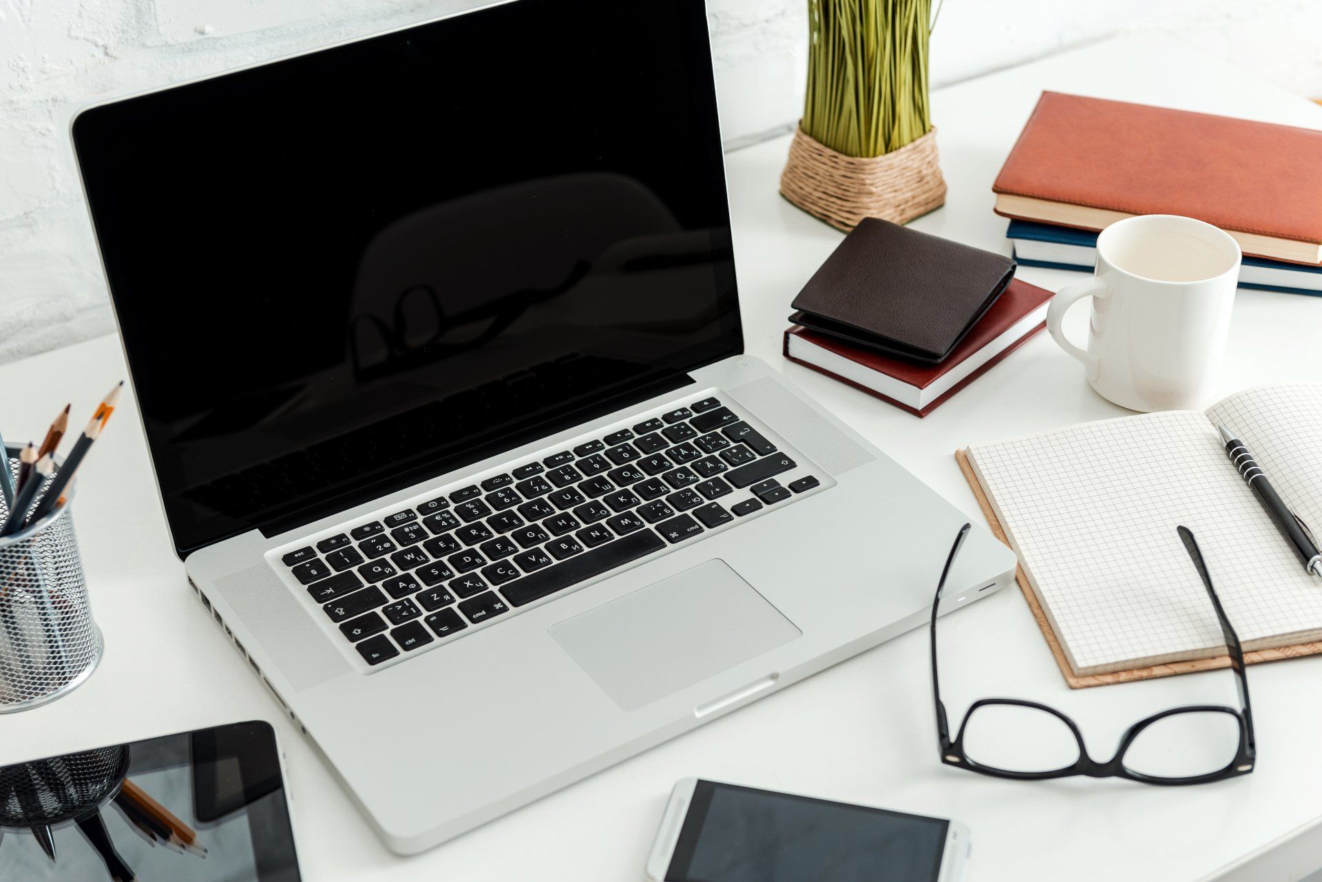 Laptop on white desk with accessories: notepad, glasses, mug, pens, and books.