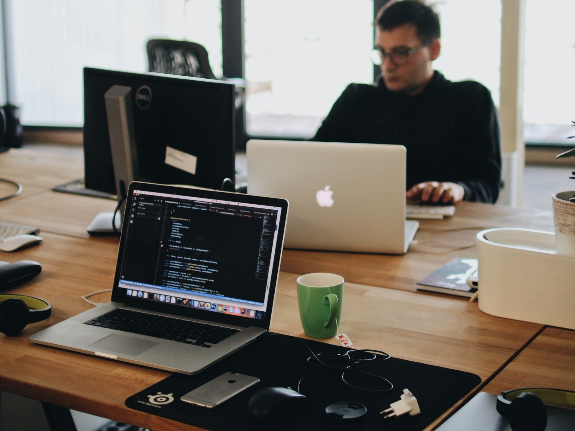 Man working on laptops in a light-filled office. One laptop displays code, another is open. Green mug nearby.