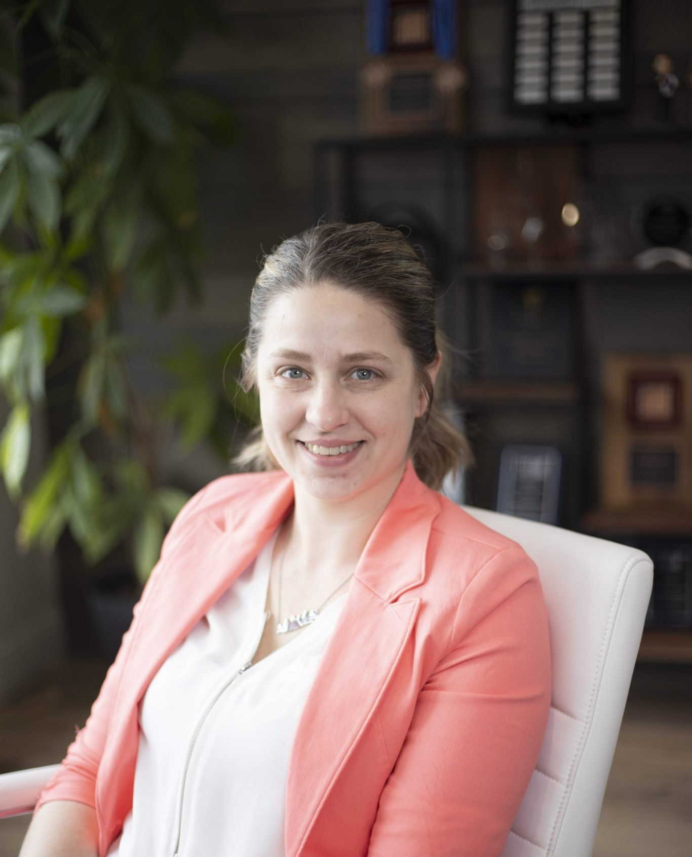 Woman wearing a coral blazer smiles, seated in a white chair. Background includes plants and a wooden shelf.