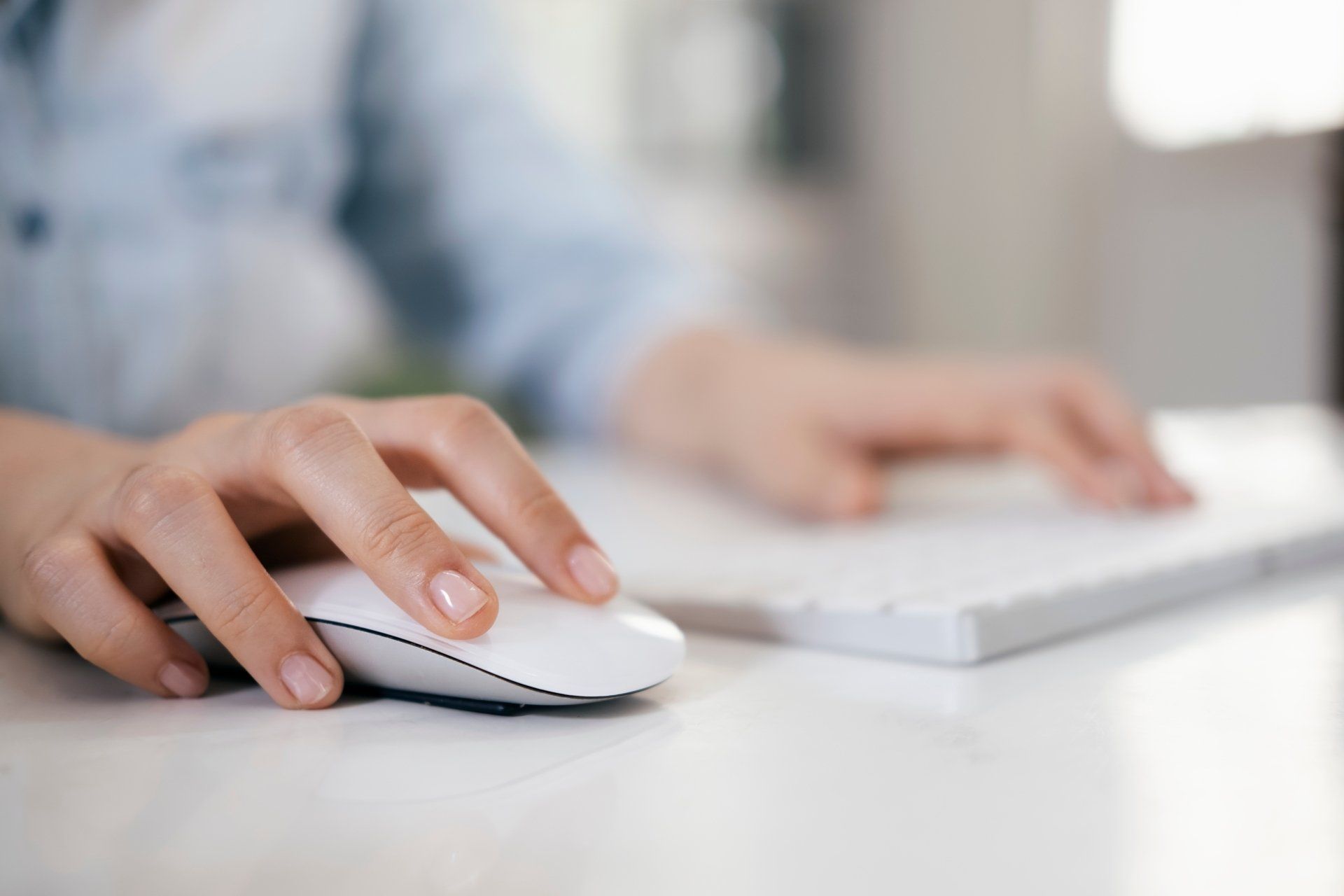 Woman's hand on a white computer mouse, with a keyboard in the background, on a white desk.