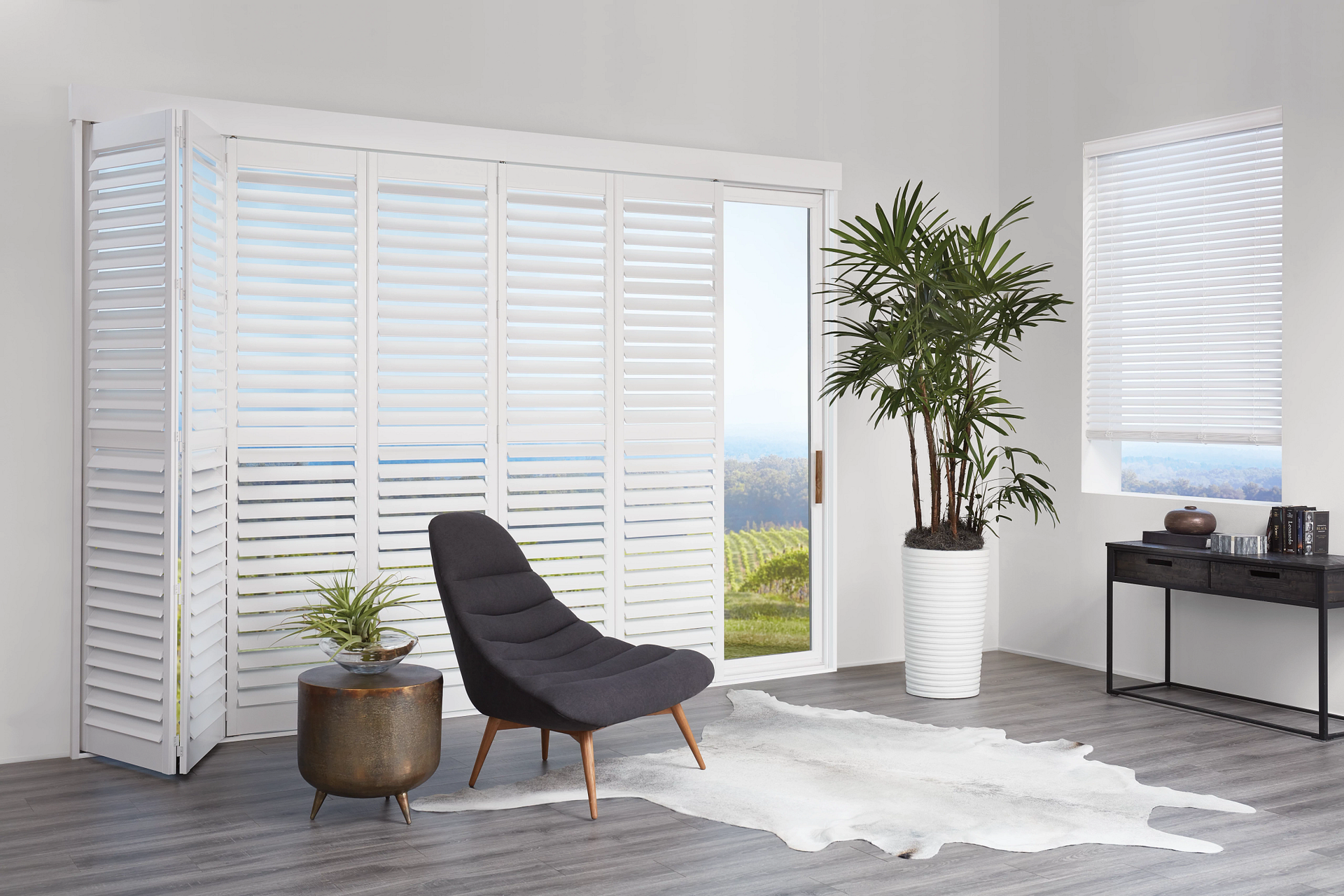 A modern living room with folding white plantation shutters on large windows, a black lounge chair, and a potted plant.