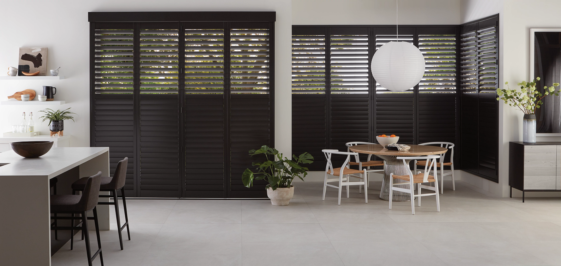 A modern dining room and kitchen area featuring dark floor-to-ceiling plantation shutters and light neutral decor.