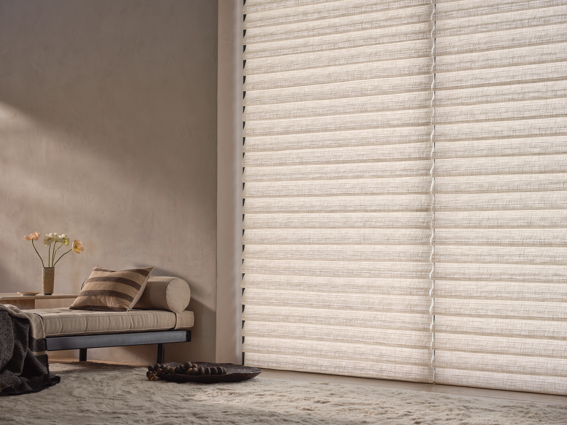 Light beige pleated window shades cover a large window next to a minimalist couch and side table in a neutral room.