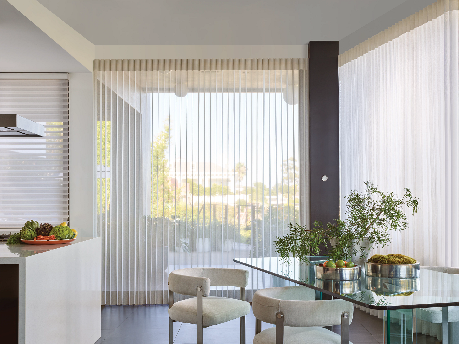 A modern dining room with a glass table, white chairs, and large windows covered by vertical, sheer blinds.