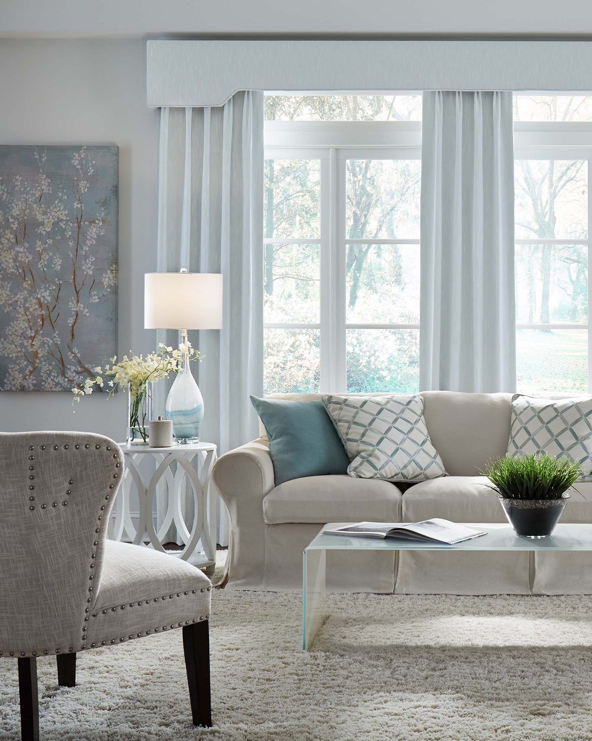 A living room with a beige sofa, patterned cushions, an armchair, a glass coffee table, and light curtains by a window.