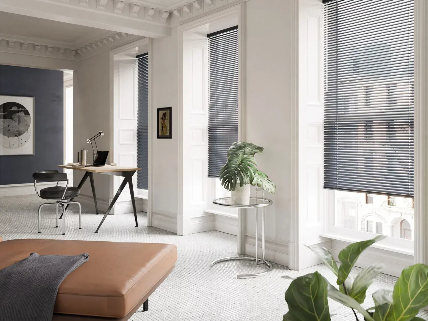 Modern room with dark blinds on tall windows, a minimalist desk, an armchair, a plant, and a brown ottoman.