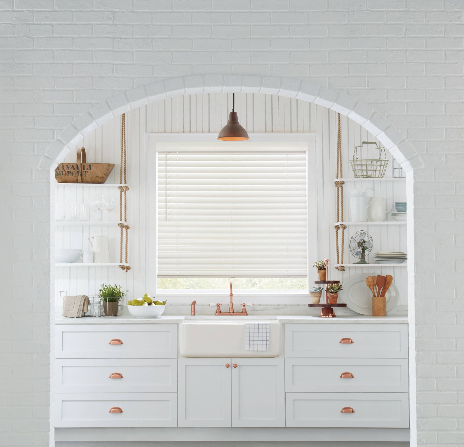 A white kitchen features a farmhouse sink, shelving hung by ropes, and a window with white blinds under an arched alcove.