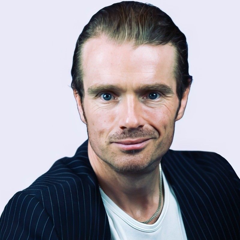 A person with short, swept-back hair wearing a pinstriped blazer over a white shirt, looking directly at the camera.