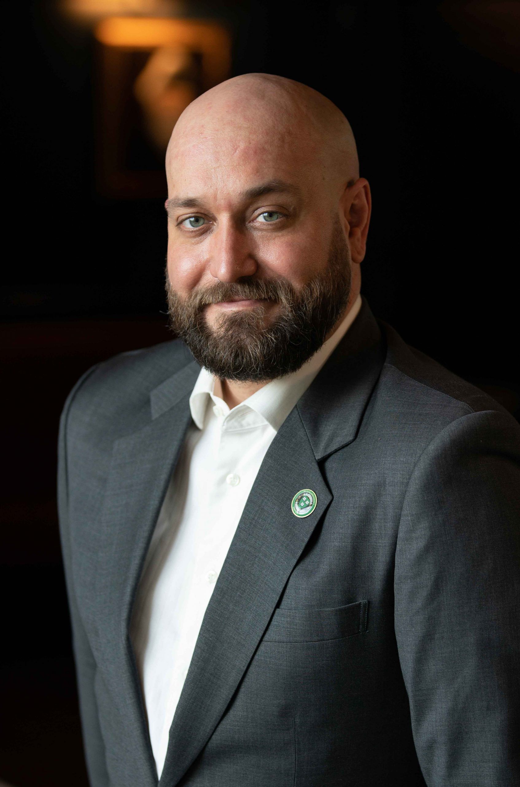A bald man with a beard, wearing a gray suit and white shirt, stands against a dark background.