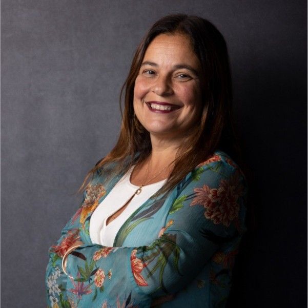 A woman in a blue floral jacket is smiling with her arms crossed.