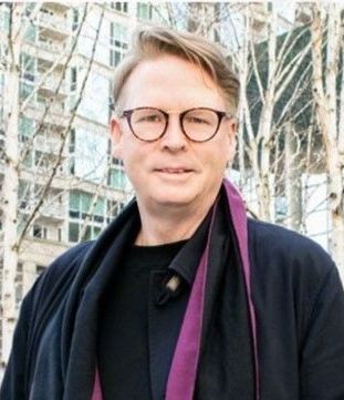 A man wearing glasses and a purple scarf is standing in front of a building.