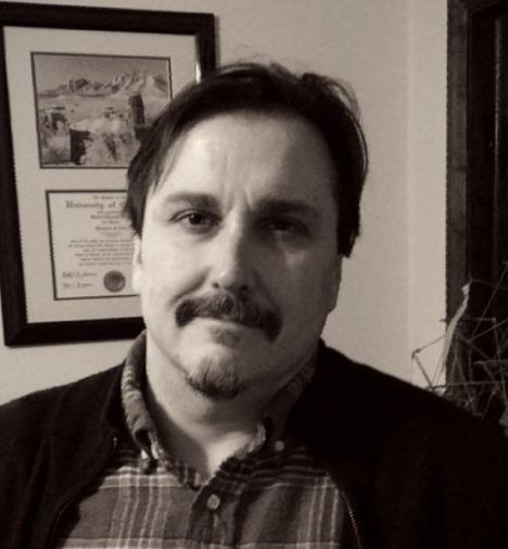 A man with a beard is wearing a plaid shirt and a black jacket