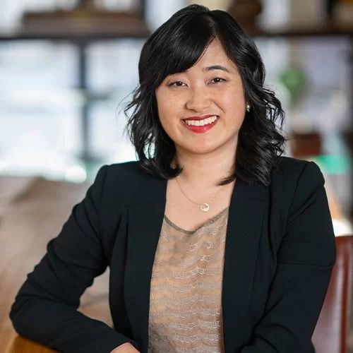 A woman in a black jacket is sitting at a table and smiling for the camera.