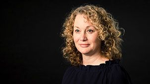 A woman with curly hair is standing in front of a black background and smiling.