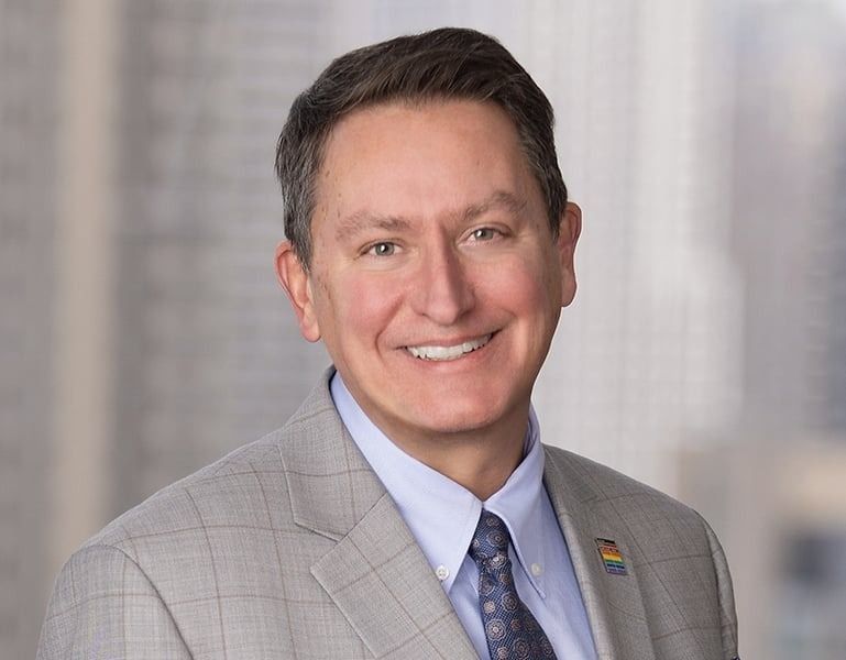 A smiling professional in a grey plaid suit jacket, light blue dress shirt, and patterned tie, set against a blurred city.