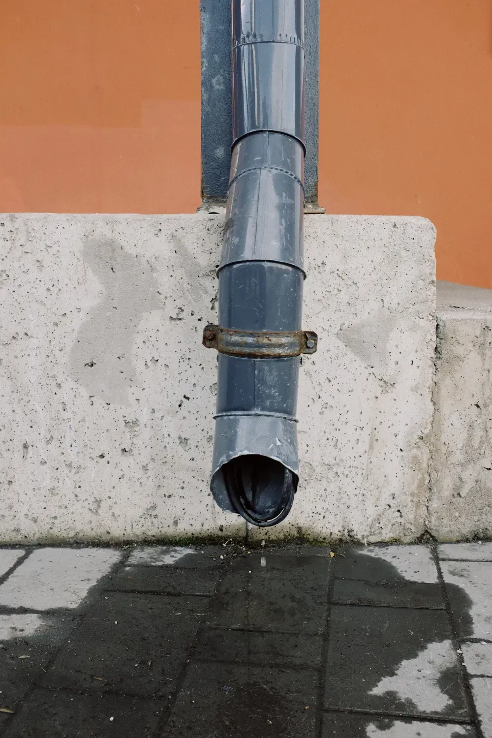 Rainwater pouring from a gray downspout onto a wet sidewalk, against an orange wall.