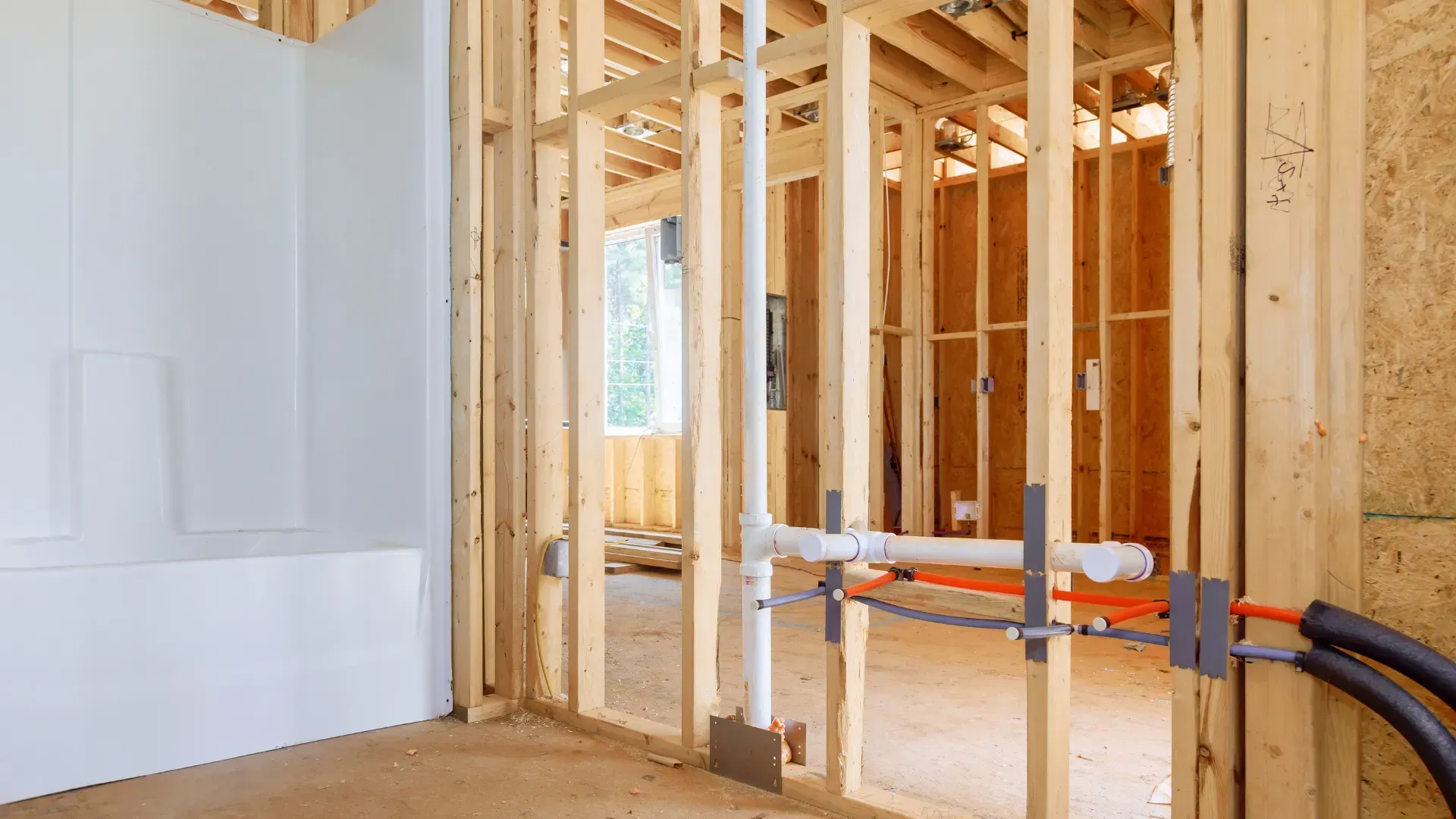 Interior of a building under construction; wooden framing, plumbing pipes, and wiring visible.