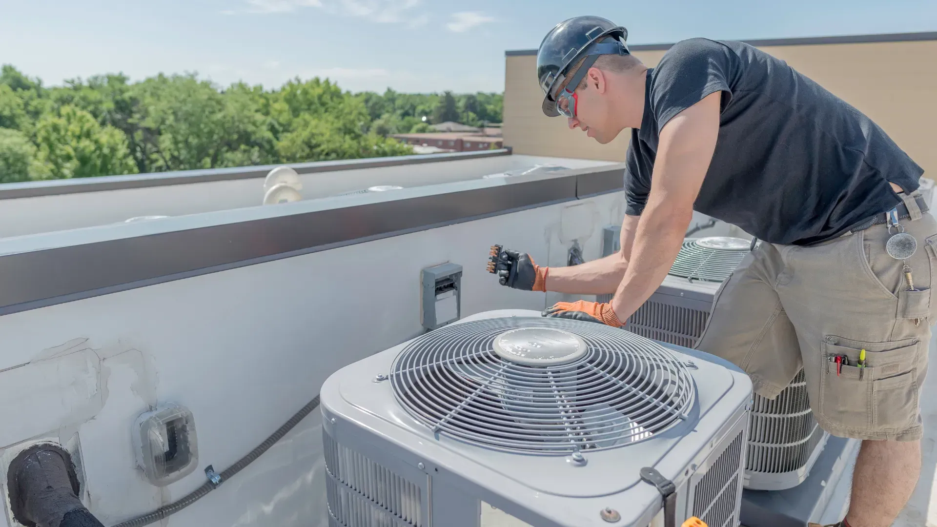 HVAC technician inspecting an air conditioning unit on a rooftop. Wearing safety gear, blue sky in background.