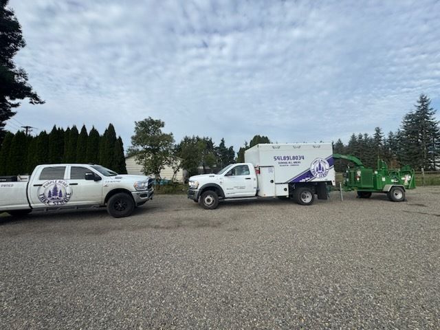 Two white work trucks and a wood chipper on gravel. Trucks have company logo.