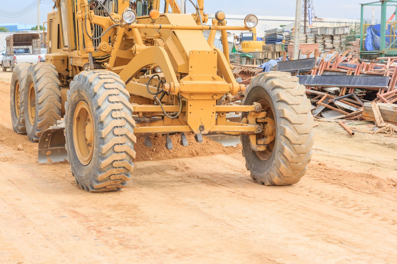 Yellow road grader leveling dirt at a construction site with debris in the background