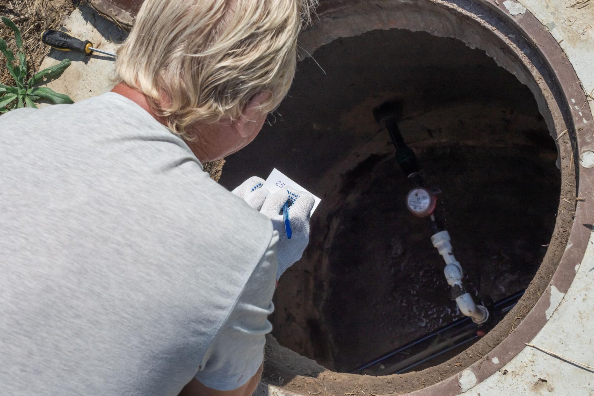 A man is looking into a manhole while holding a piece of paper.