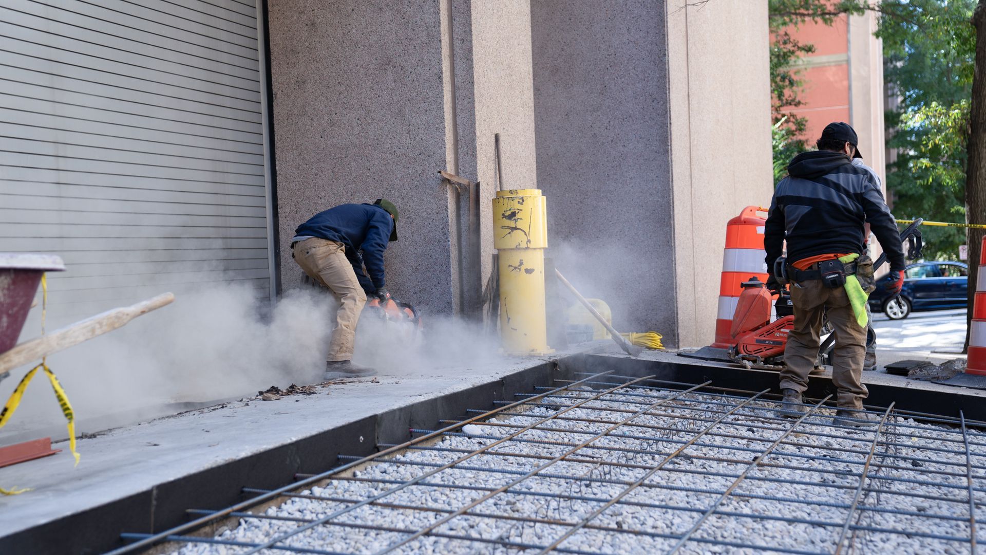 Two construction workers are working on a sidewalk in front of a building.