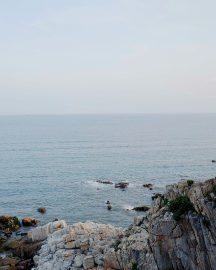 Rocky seaside cliff overlooking a calm, pale blue ocean under a hazy sky.