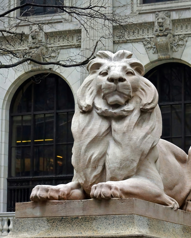 Stone lion statue in front of a building with arched windows and ornate stonework.