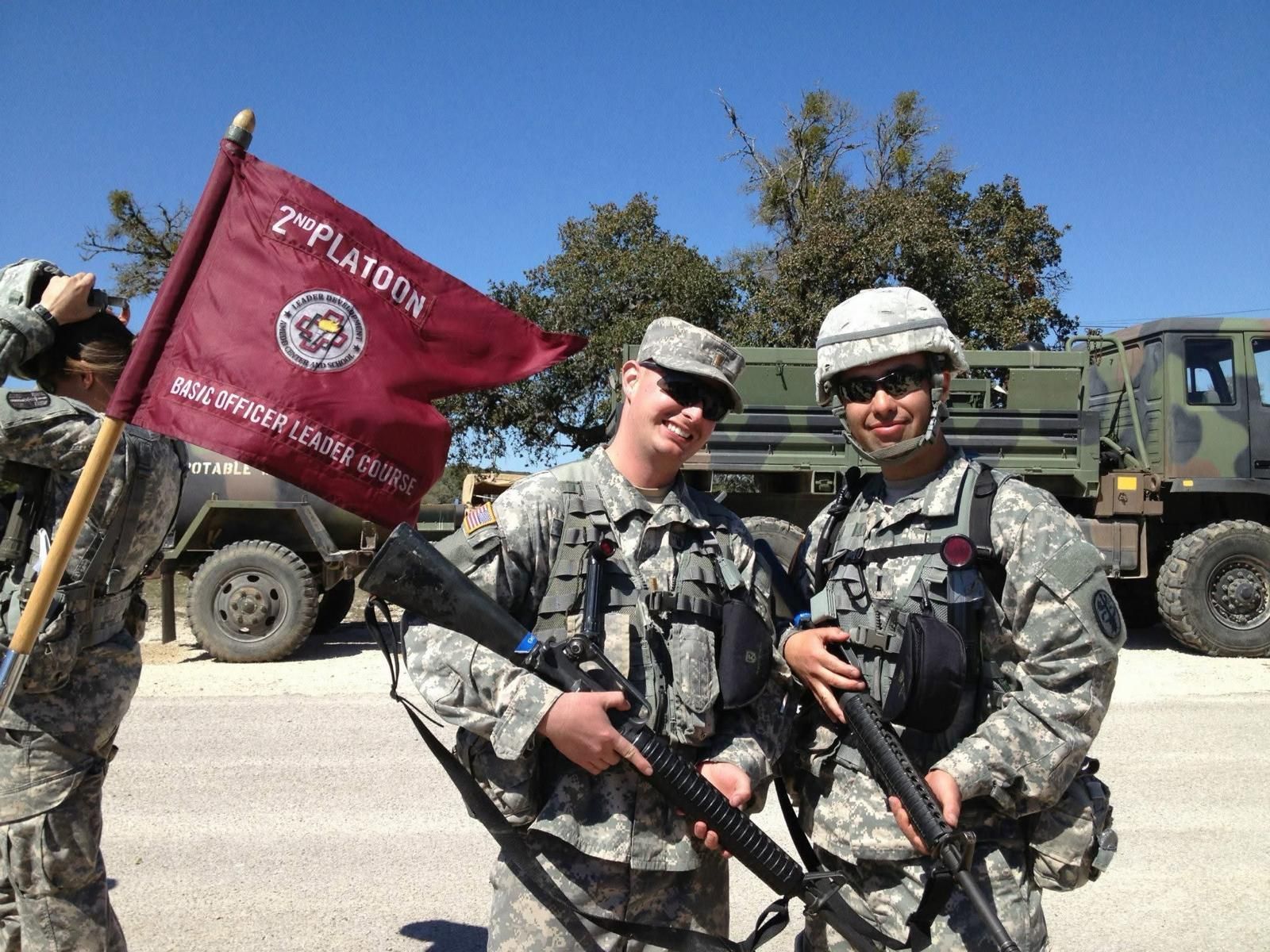 Two soldiers in camouflage hold rifles beside a red military flag and vehicle outdoors.