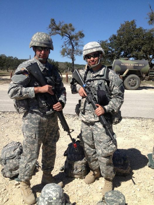 Two soldiers in camouflage uniforms and helmets stand holding rifles beside a military vehicle.