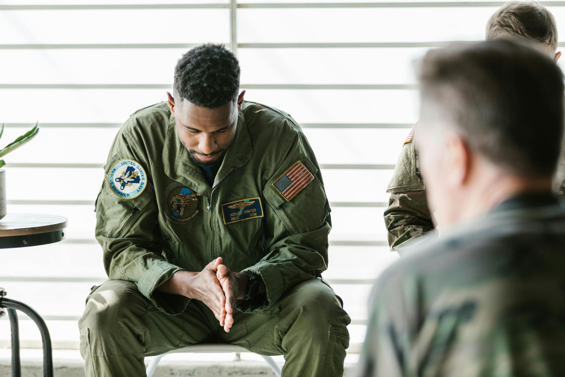 Man in green flight suit sits with head bowed in a bright room during a group meeting
