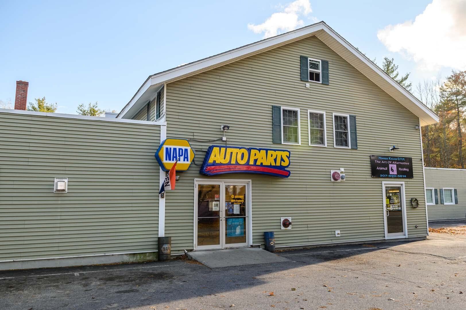 A light green, multi-level building with a NAPA Auto Parts sign, entrance doors, and gravel parking lot.